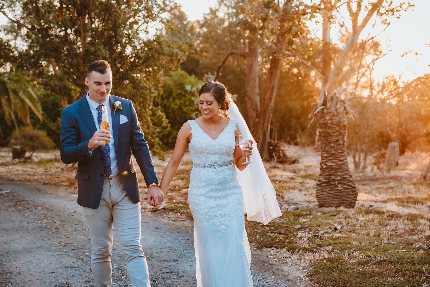 A couple in wedding attire walking together on a dirt path at sunset while holding hands and drinks.
