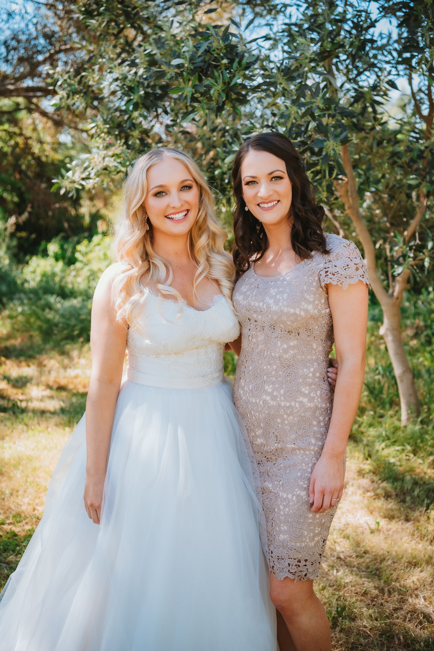 Two women pose outdoors in formal dresses - one in a flowing white gown and one in a beige lace cocktail dress.
