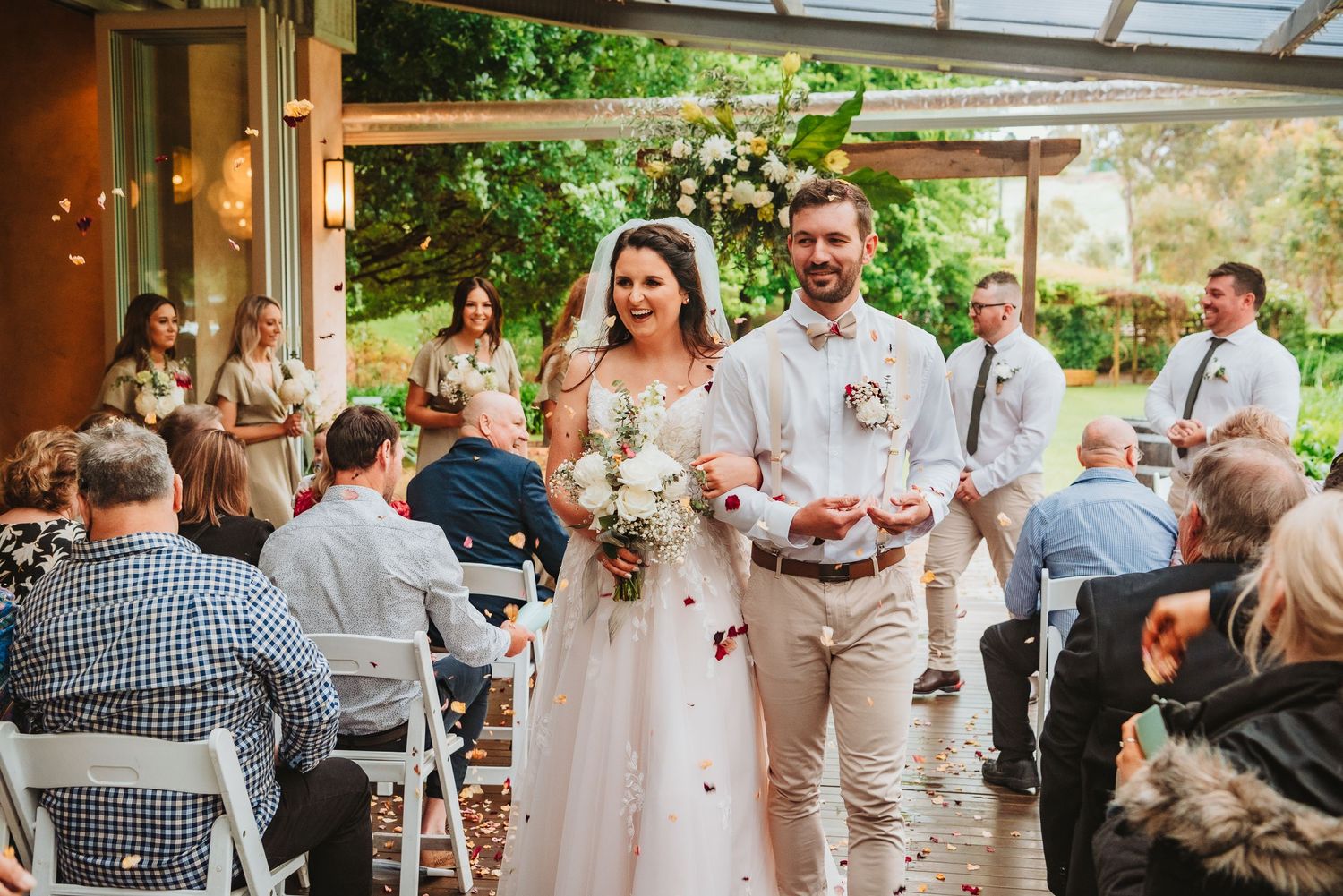 A joyous wedding ceremony outdoors under a covered patio with guests seated in white chairs.