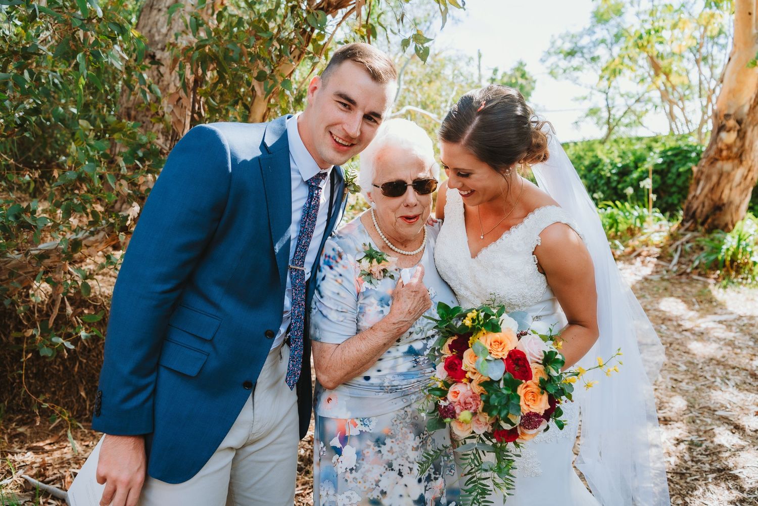 A wedding couple shares a joyful moment with an elderly guest in a garden setting while holding a vibrant floral bouquet.