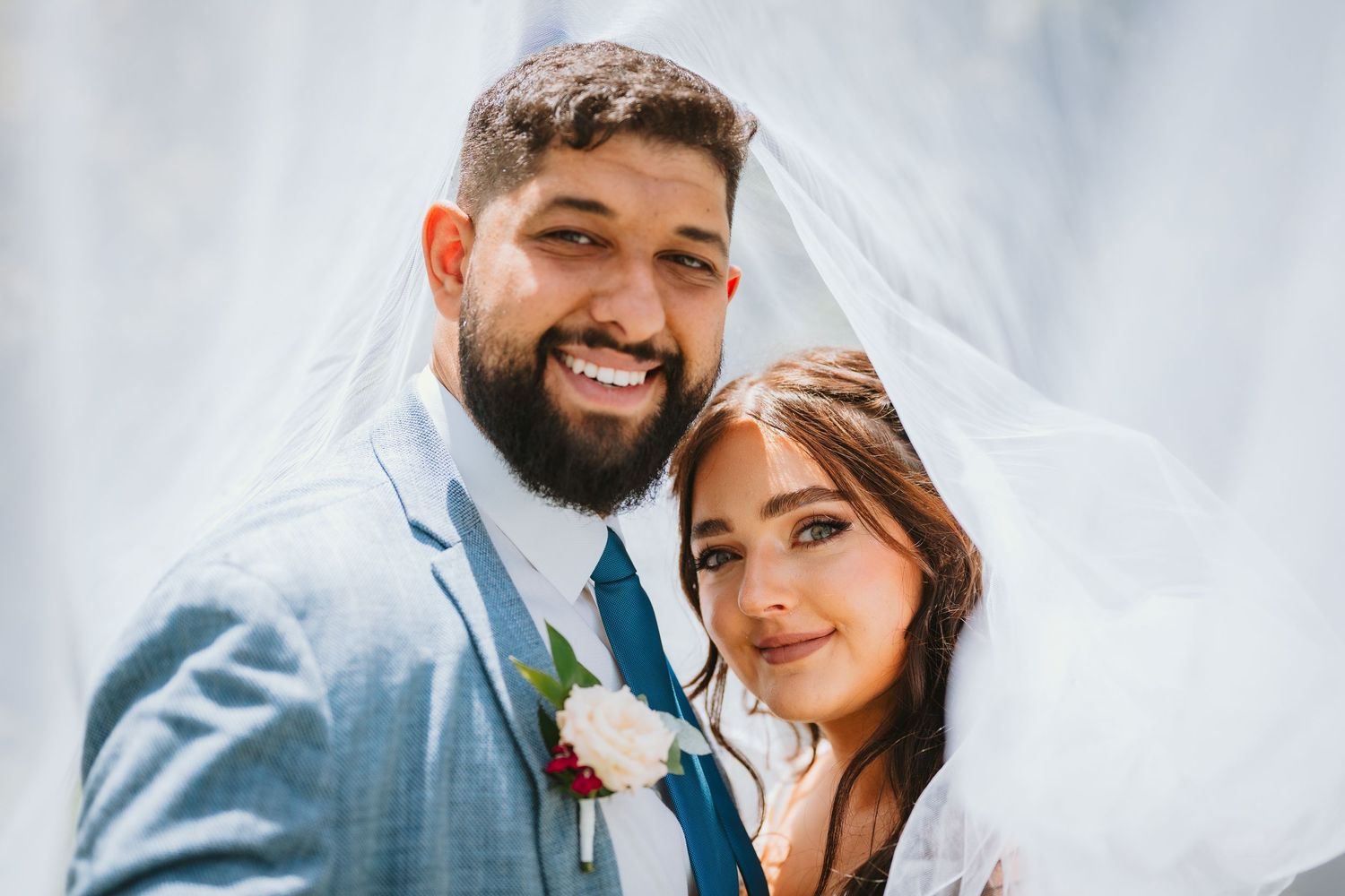 Newlywed couple share an intimate moment together outdoors on their wedding day wearing light blue suit and white gown.