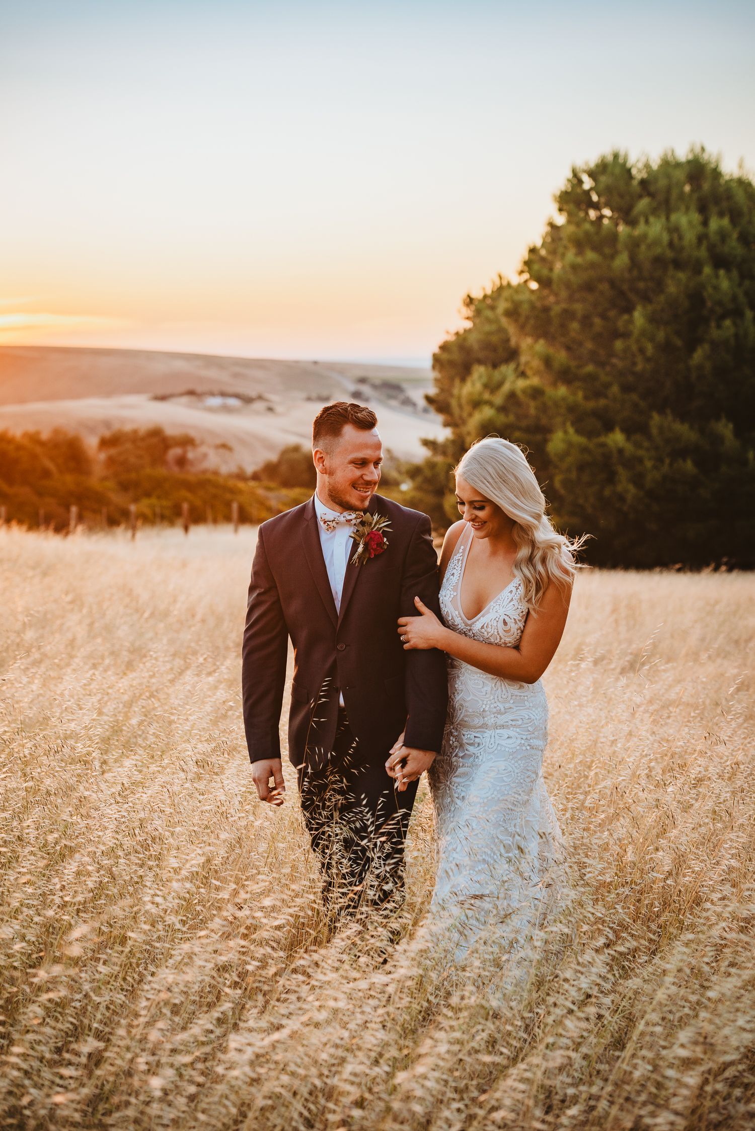 A couple walks together through a golden wheat field at sunset with mountain views in the background.