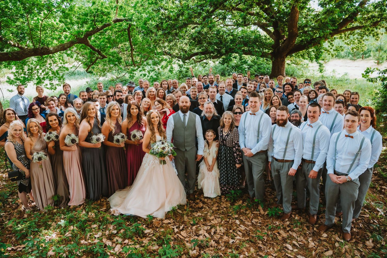 Large wedding party poses together under a sprawling tree with bridesmaids in brown dresses and groomsmen in gray suits.