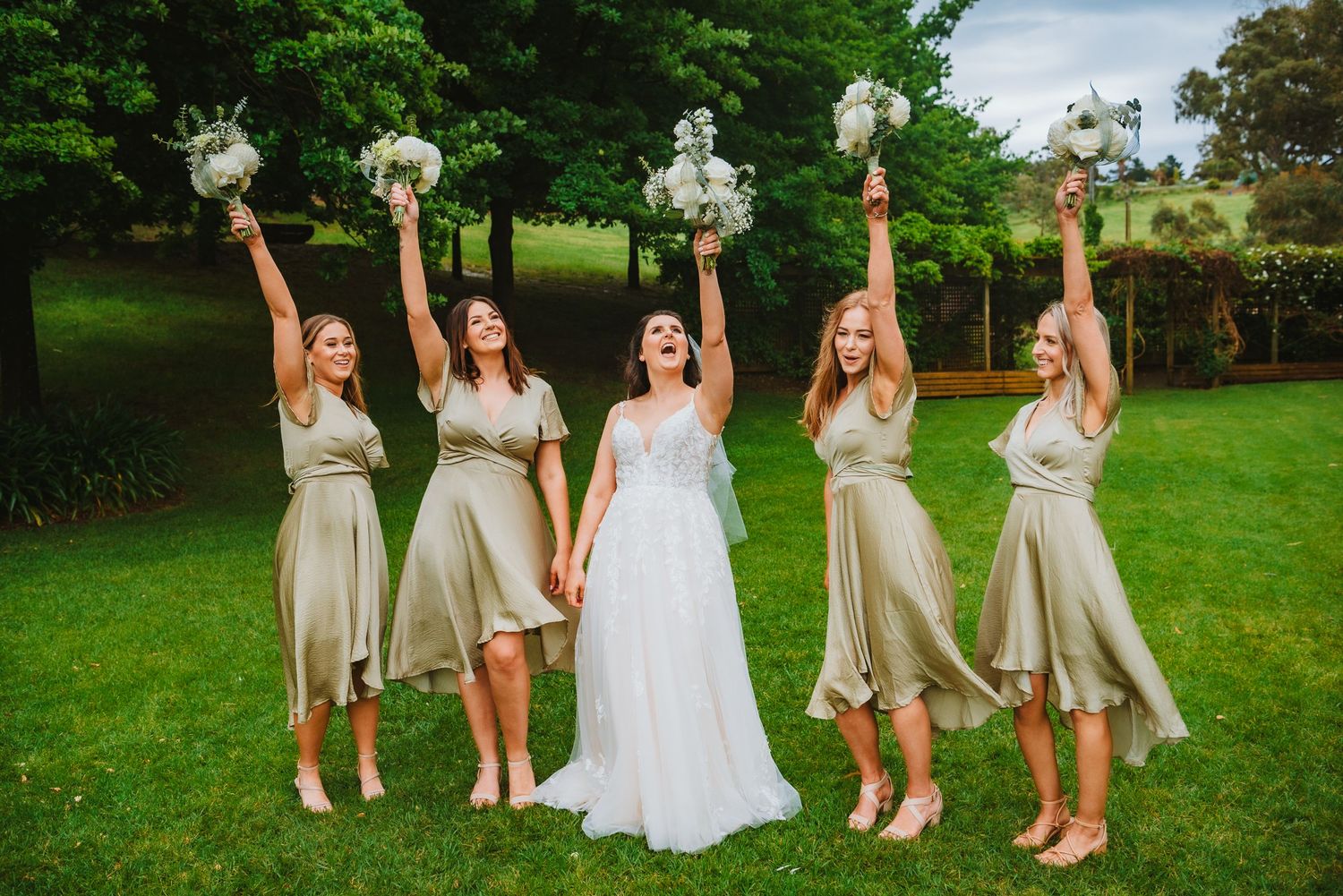 Bridesmaids in beige dresses and white bouquets celebrate joyfully with bride in white gown on green lawn.