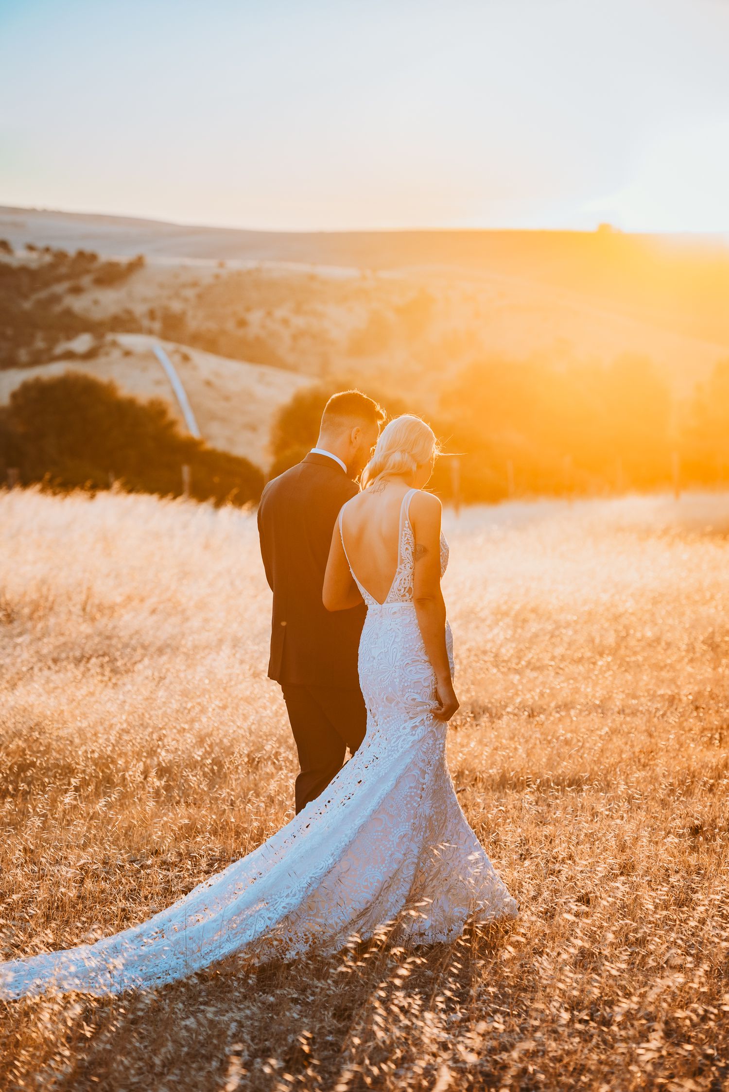 A couple stands together on a golden hilltop during sunset as the bride's lace wedding dress trails behind them.