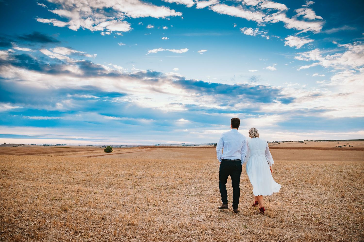 Two people hold hands while walking in a vast desert landscape under dramatic blue skies with white clouds.