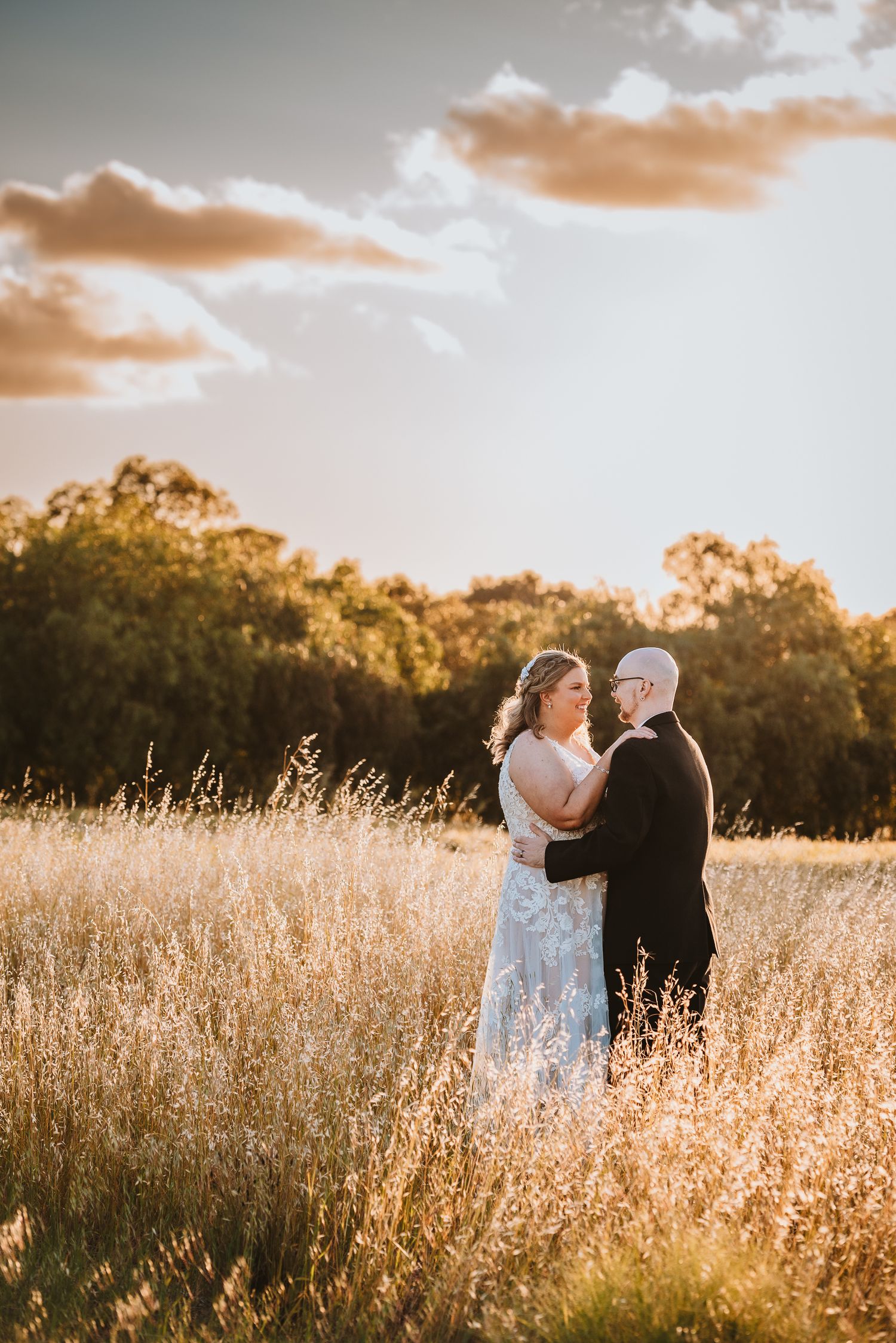 A couple embraces in a golden wheat field at sunset with trees and dreamy clouds in the background.