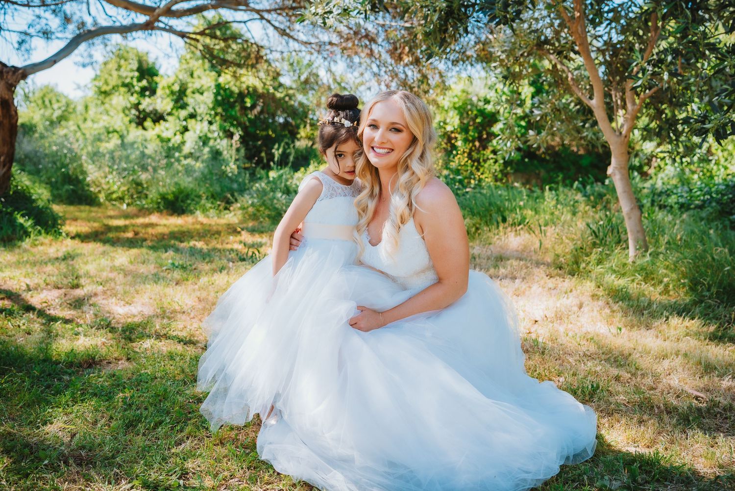 Two people in white wedding dresses embrace while sitting together in a sunny garden setting.