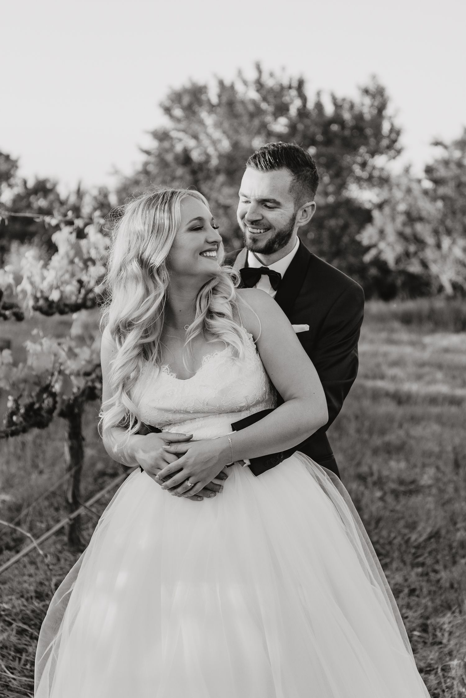 A black and white wedding photo captured outdoors shows a joyful couple embracing in wedding attire against a natural backdrop.