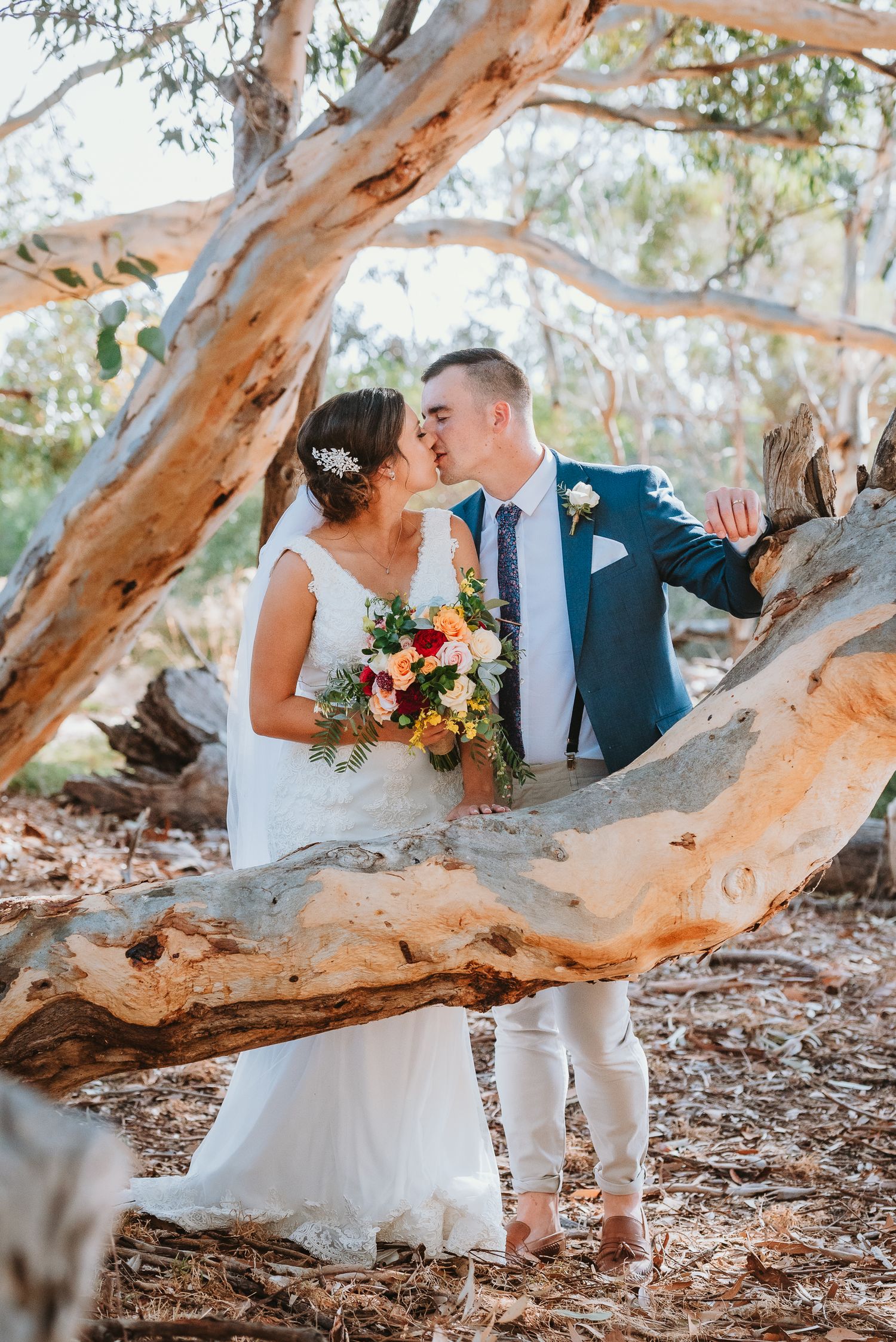 A romantic wedding photo captured under a twisted eucalyptus tree showing a navy suited groom and bride in white with vibrant bouquet.