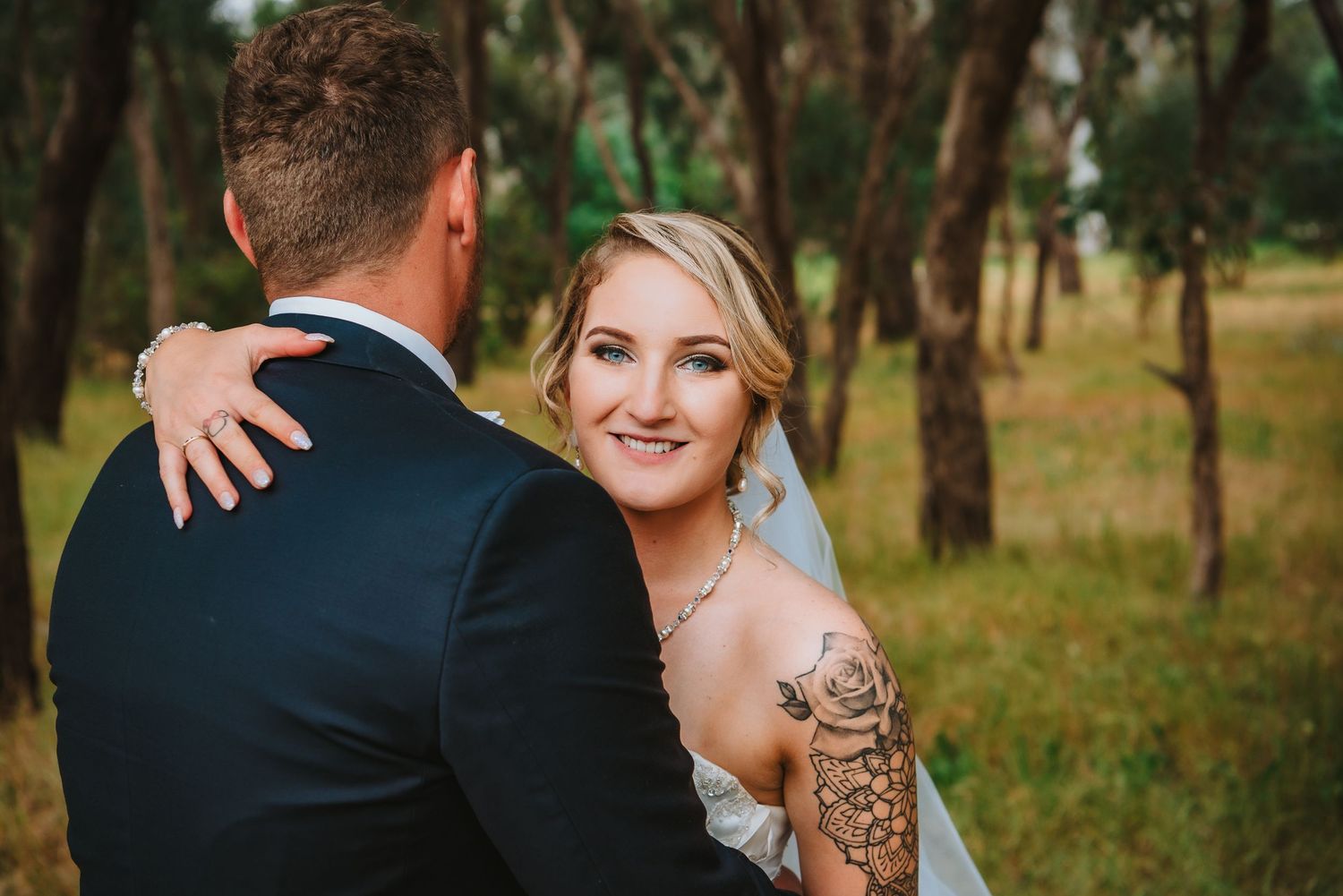 A bride with a shoulder tattoo gazes lovingly at her groom in a wooded outdoor setting at golden hour.