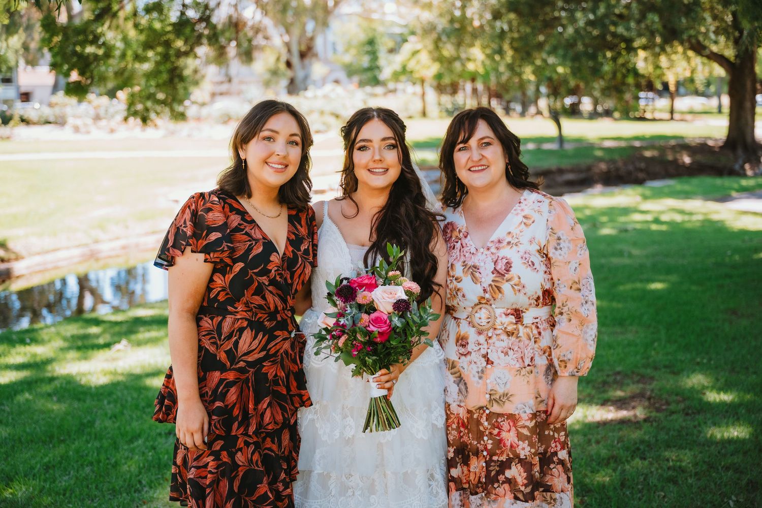 Three women pose together outdoors on a sunny day, wearing floral dresses with one in white holding a colorful bouquet.