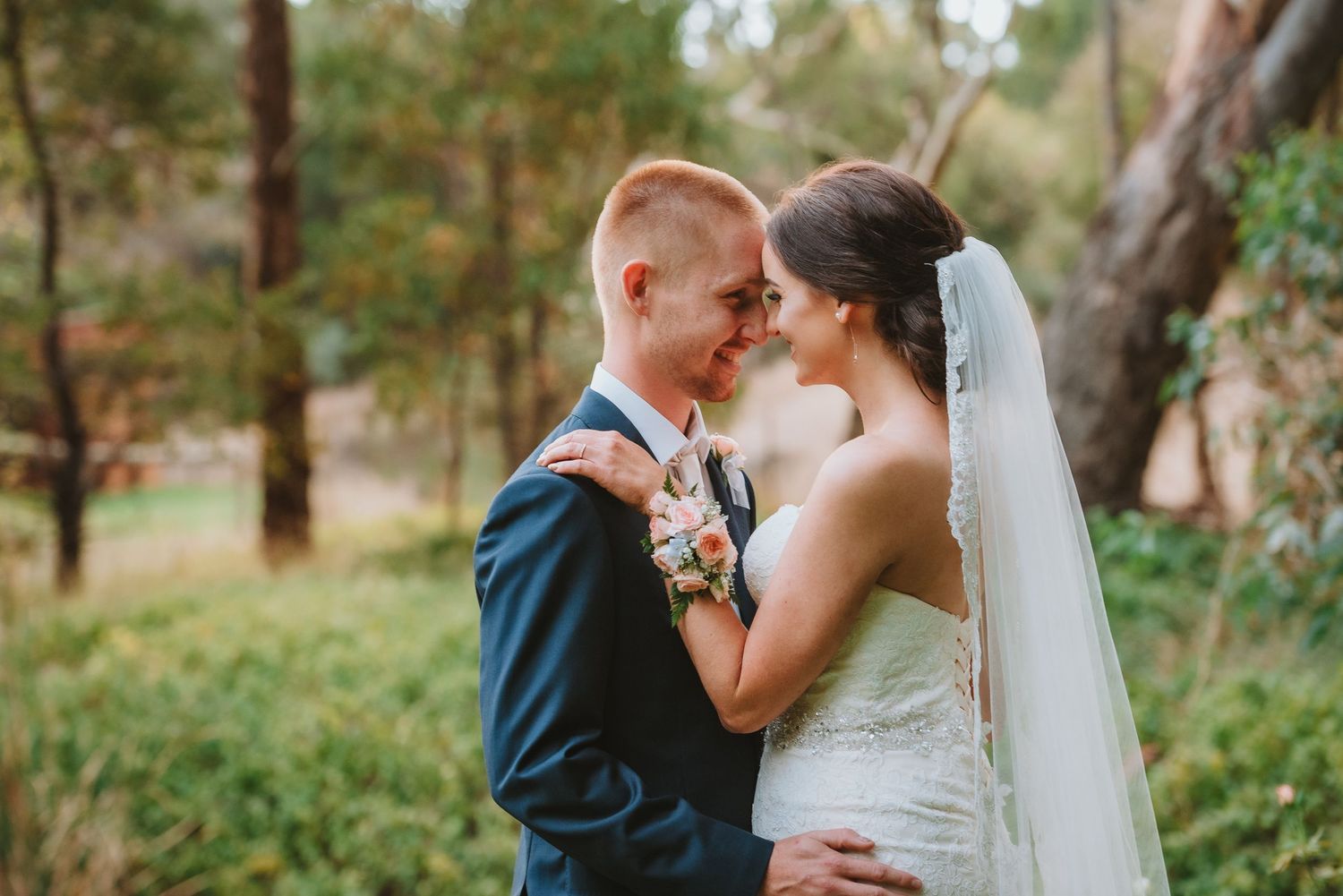 A bride in a white dress and groom in navy suit share an intimate moment outdoors among trees during their wedding.