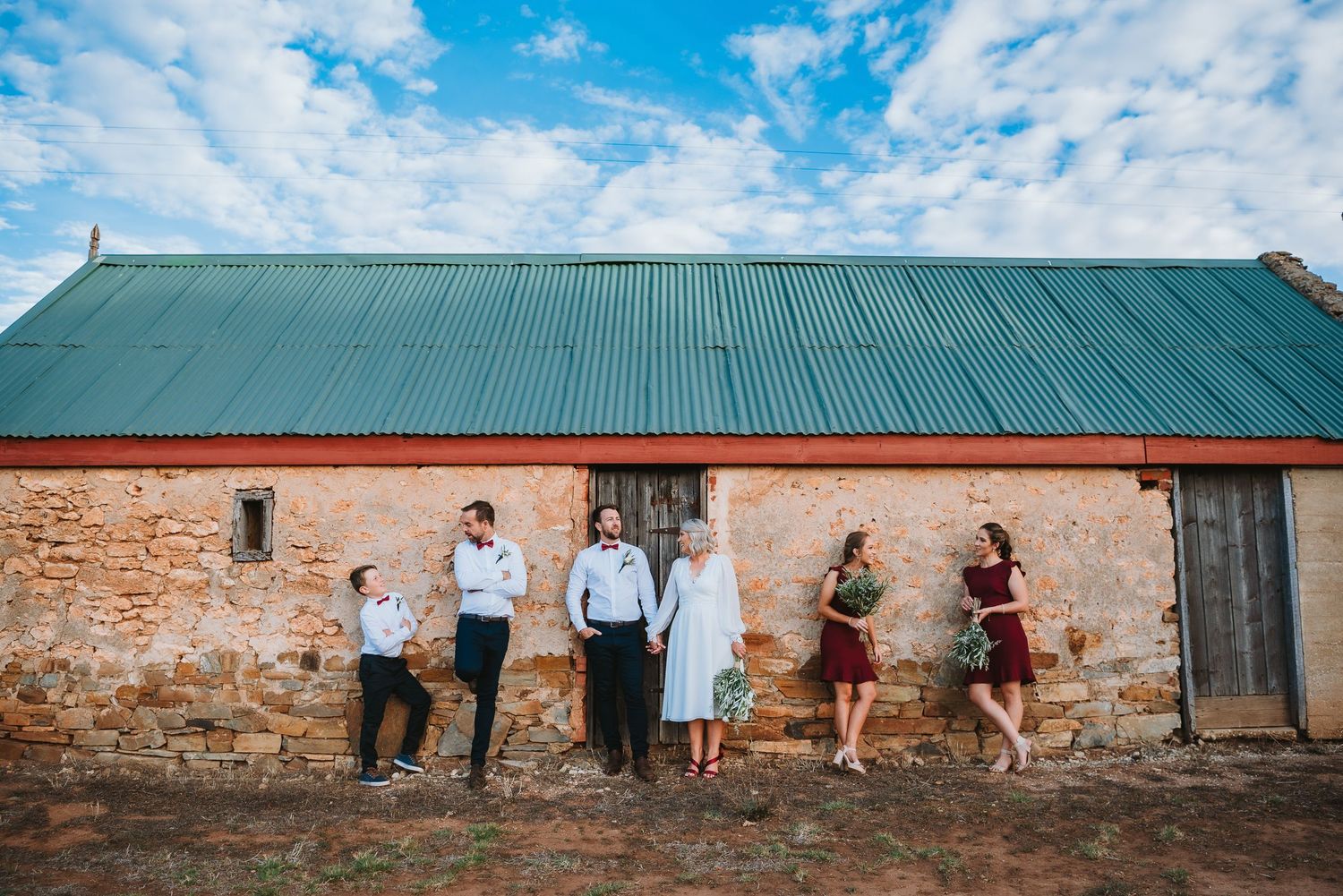 A group of people stand along a rustic brick wall with a green metal roof under a blue cloudy sky.