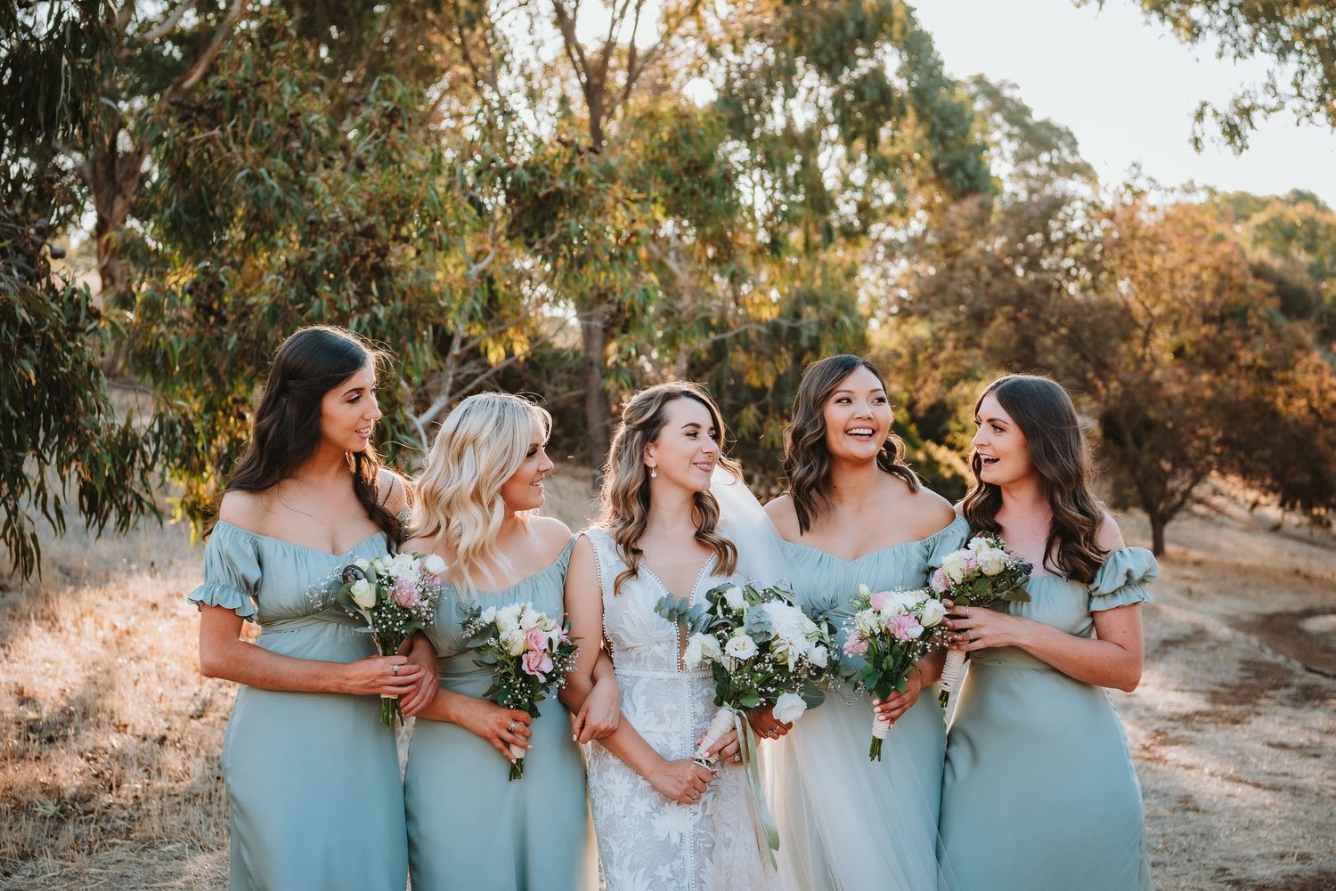 Bridal party in elegant mint blue dresses holding floral bouquets smile together during outdoor photo session.