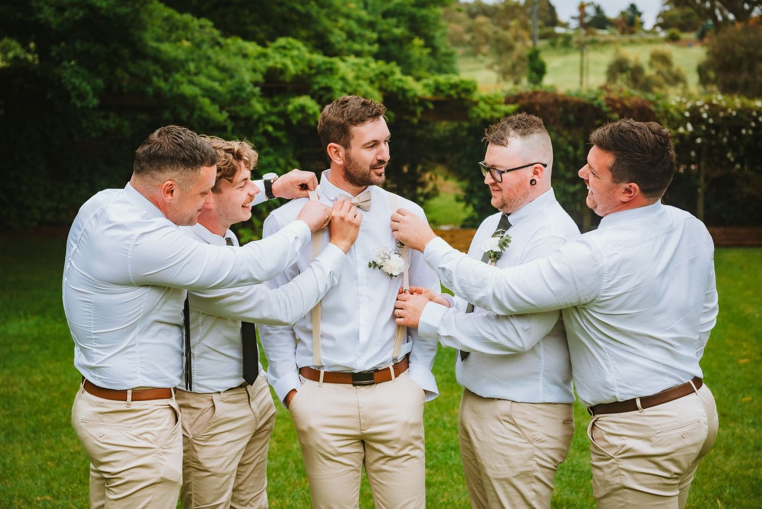 Groomsmen in matching khaki pants and white shirts toast with drinks during outdoor wedding celebration.