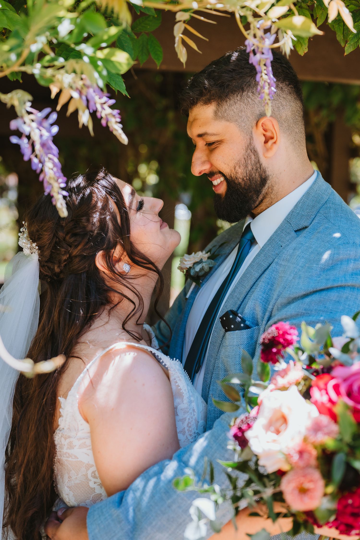 A couple share a romantic moment under blooming wisteria with colorful wedding flowers in the foreground.