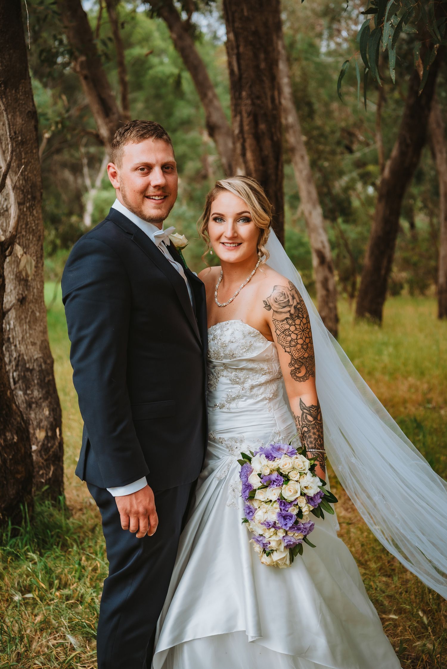 An outdoor wedding photo with a couple in formal attire, featuring a white gown, dark suit, and purple floral bouquet.
