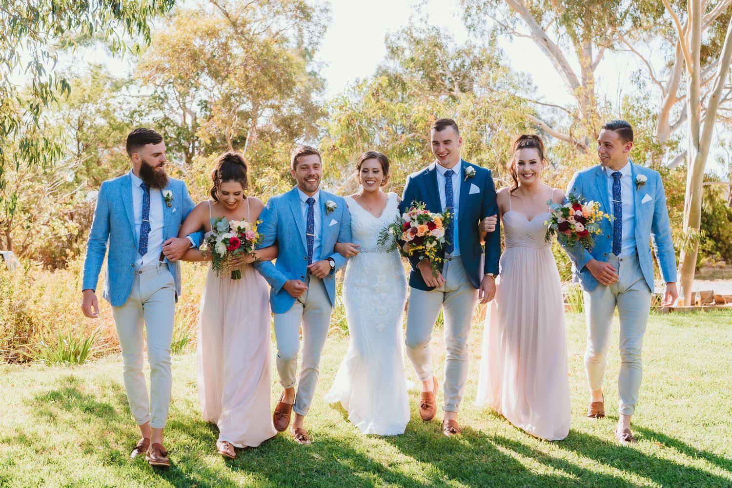 Wedding party wearing light blue jackets and pink dresses poses together outdoors on sunny day with bouquets.