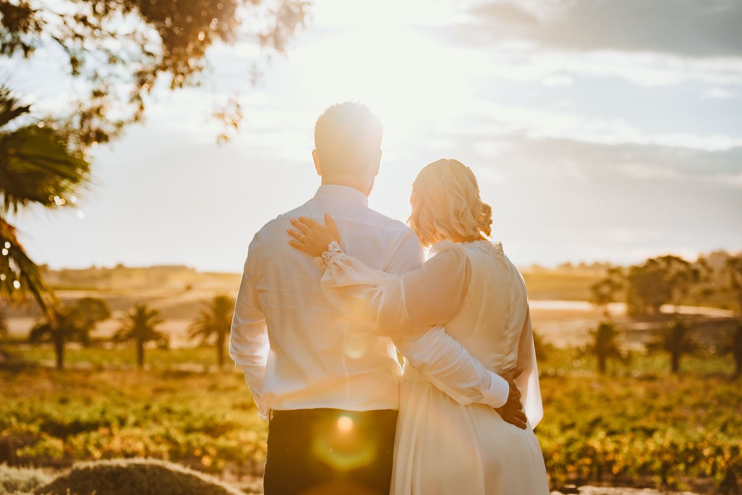 A couple in white clothing embrace while watching the sunset over a scenic rural landscape with golden light.