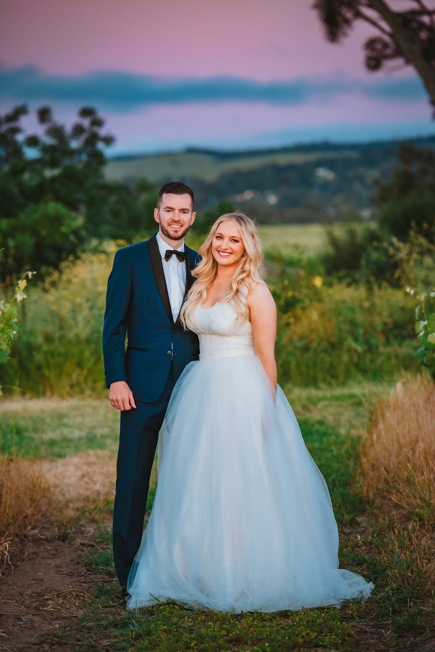 A couple celebrates their wedding day outdoors at sunset, with the bride in white and groom in navy blue against rolling hills.