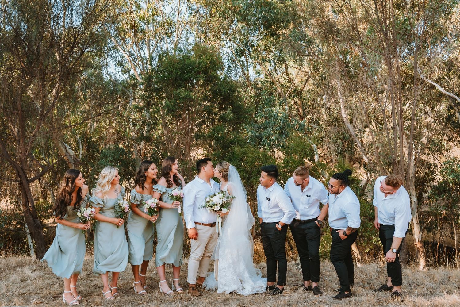 Wedding party in light blue and white attire pose casually together against a backdrop of eucalyptus trees.