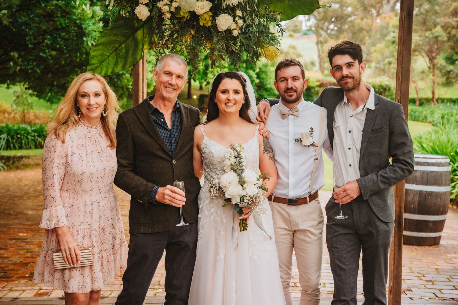 A small wedding group poses together under a floral arch in an outdoor garden venue with wine barrels.