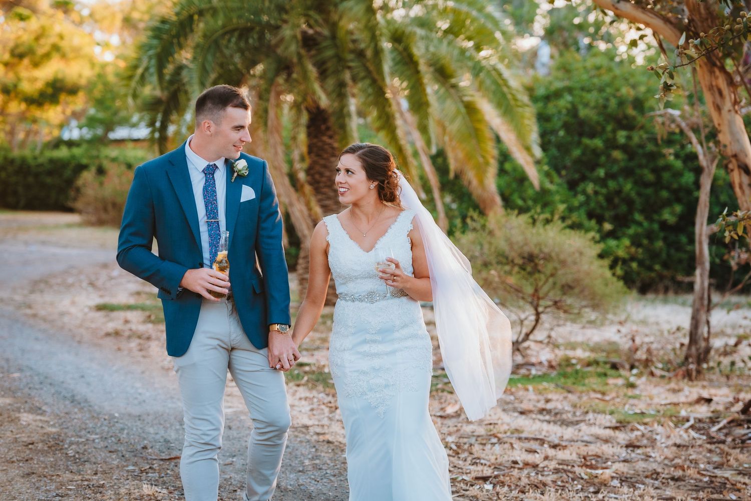 Joyful couple shares a laugh while walking down a palm-lined path in elegant wedding attire during golden hour.