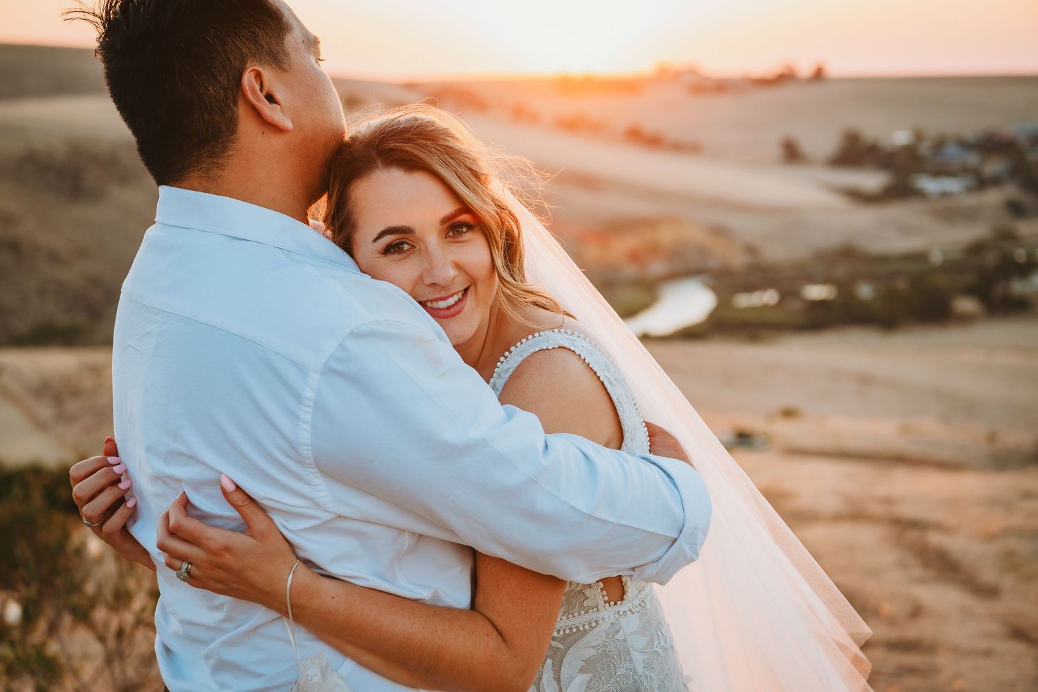 A couple embraces on a cliff at sunset, the bride wearing a white veil as golden light illuminates the coastal landscape.
