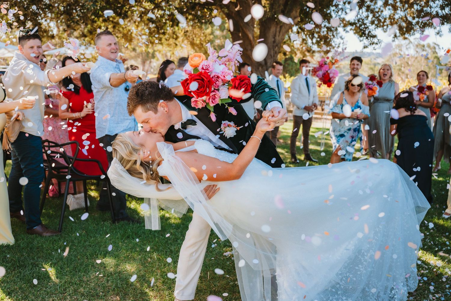 A couple shares a romantic dip kiss at their outdoor wedding ceremony while guests throw flower petals in celebration.