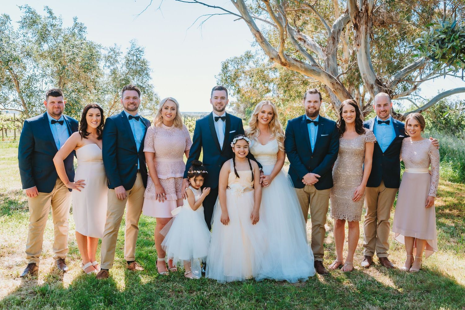 A wedding party in coordinated pink and blue outfits poses together outdoors beneath eucalyptus trees.