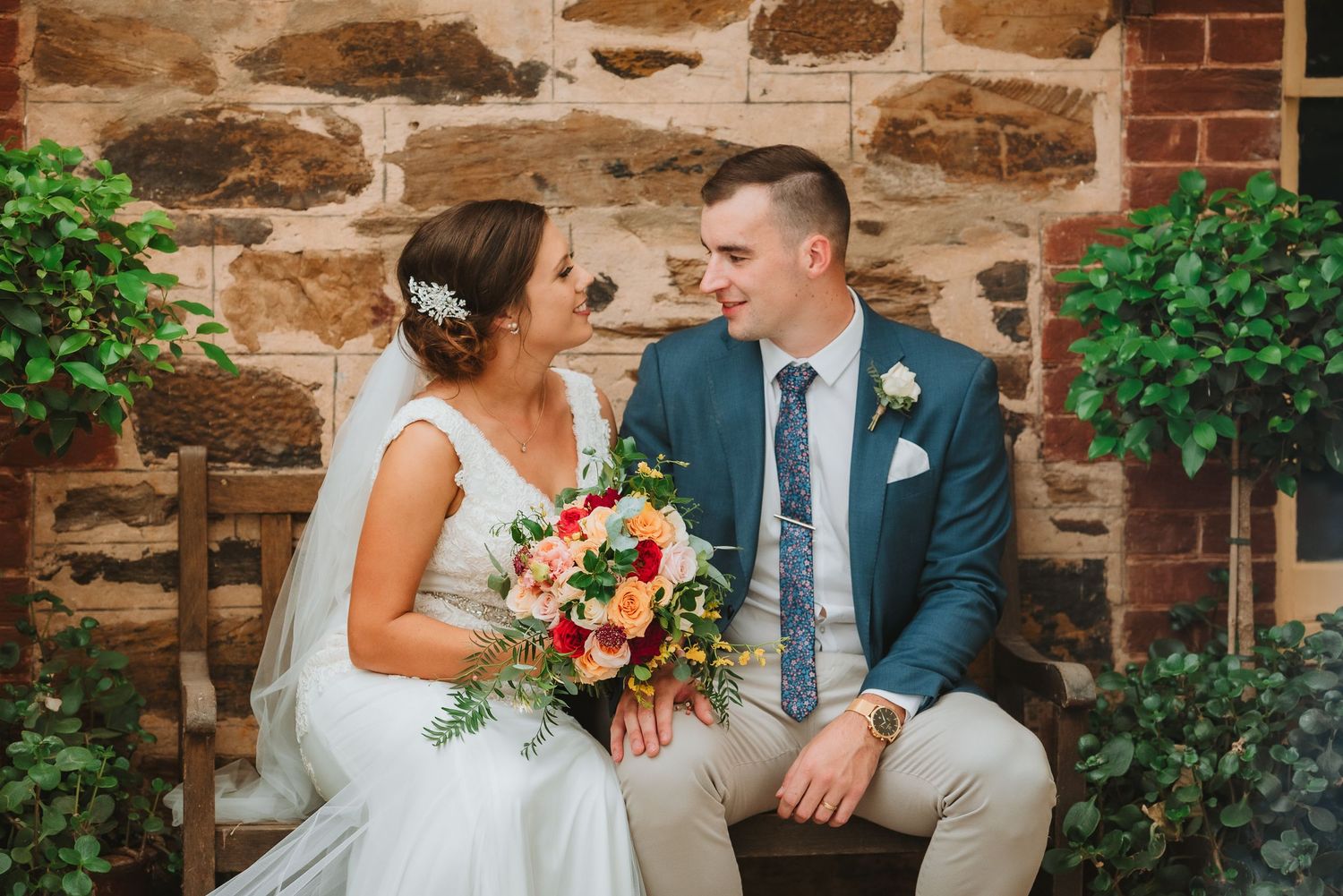 Bride wearing white dress and groom in blue suit sit together on stone wall holding bright floral bouquet.