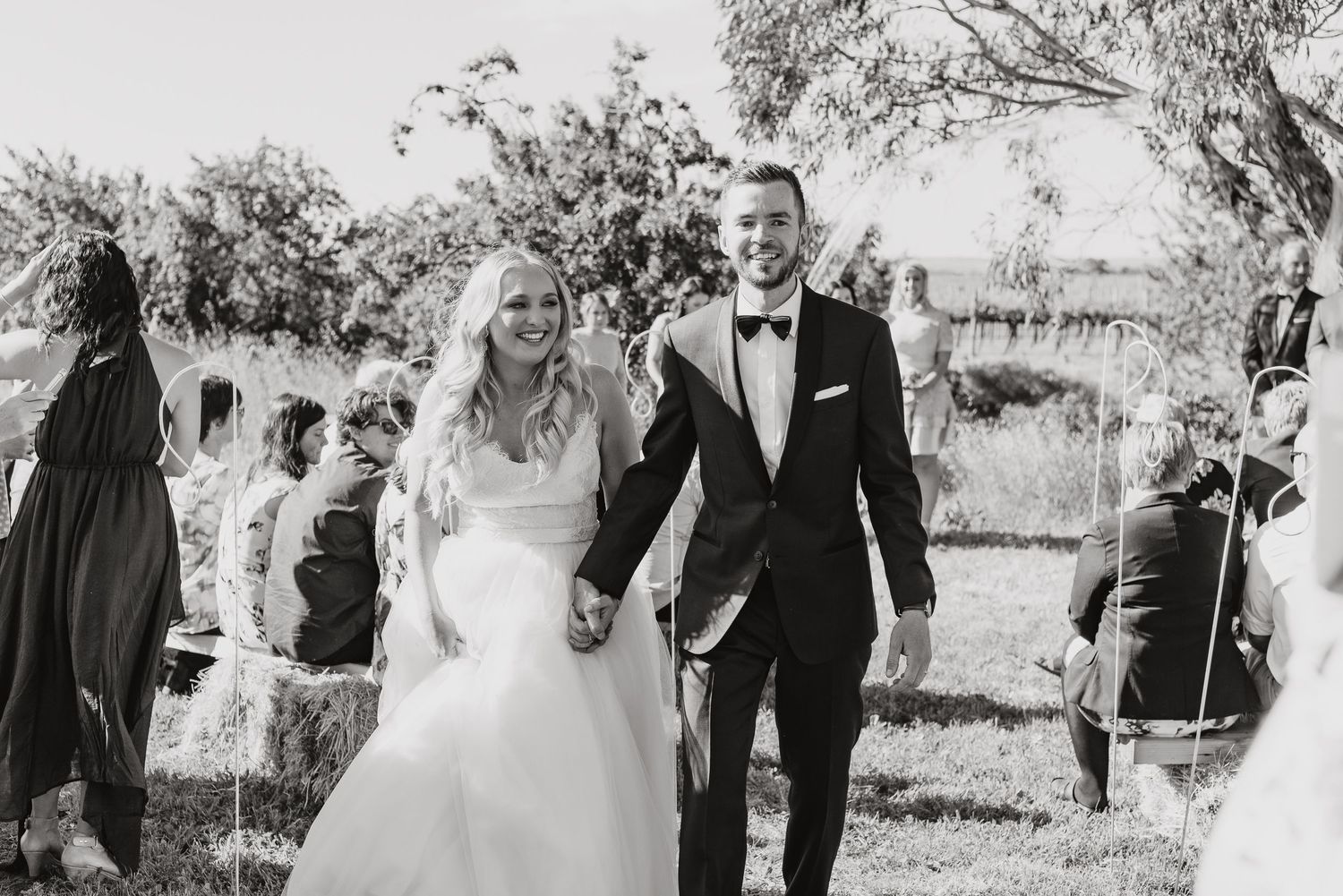 A black and white outdoor wedding moment showing a joyful couple walking hand in hand through a garden setting.