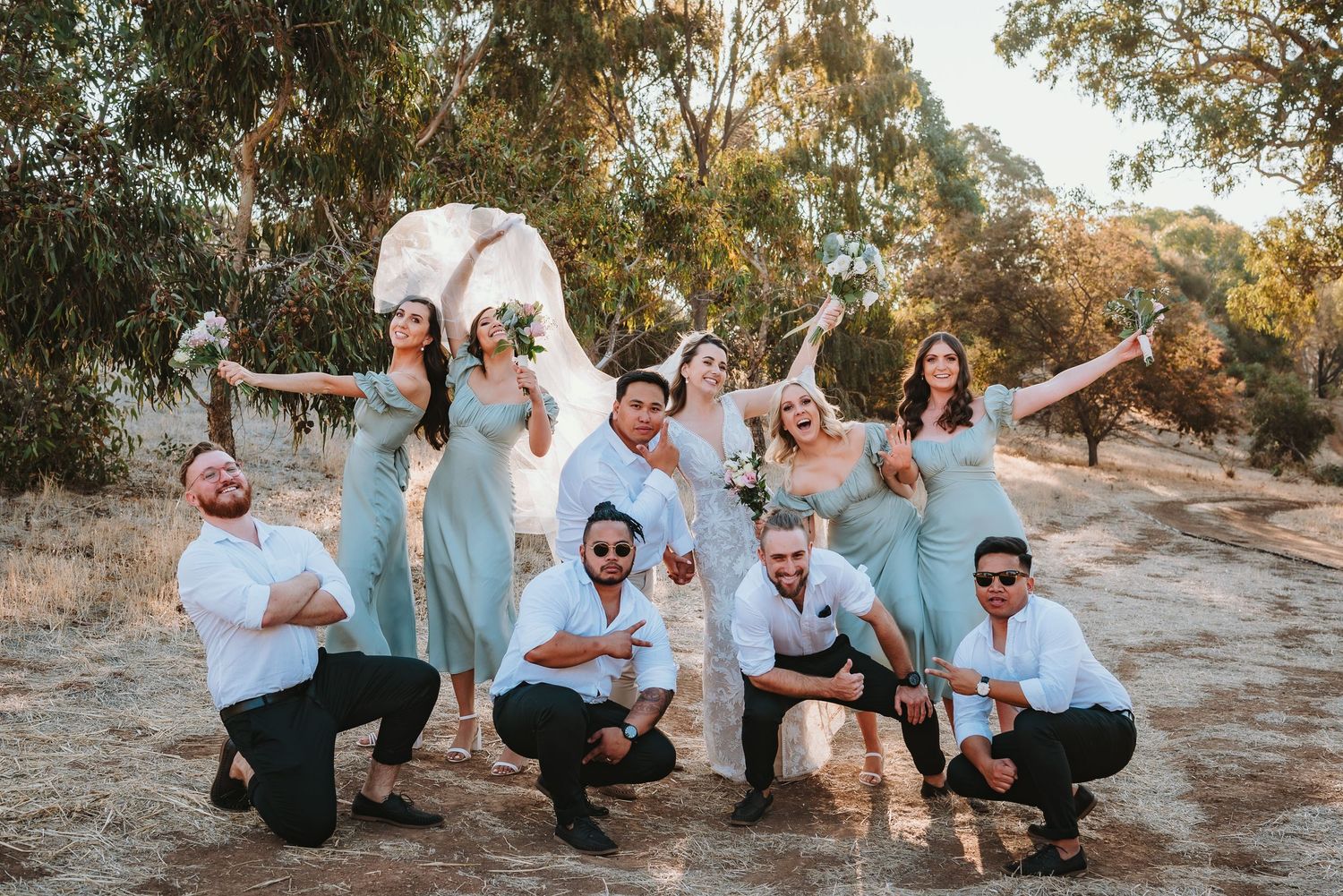 Wedding party in light blue dresses and white shirts poses playfully together outdoors in a natural setting at sunset.