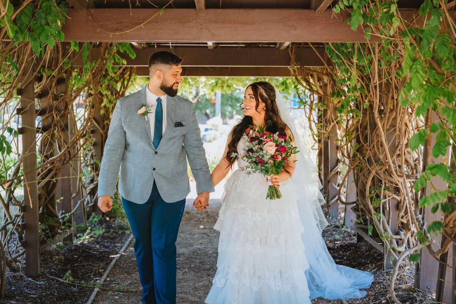 A couple shares a romantic moment under a vine-covered pergola on their wedding day, with sunlight filtering through the leaves.