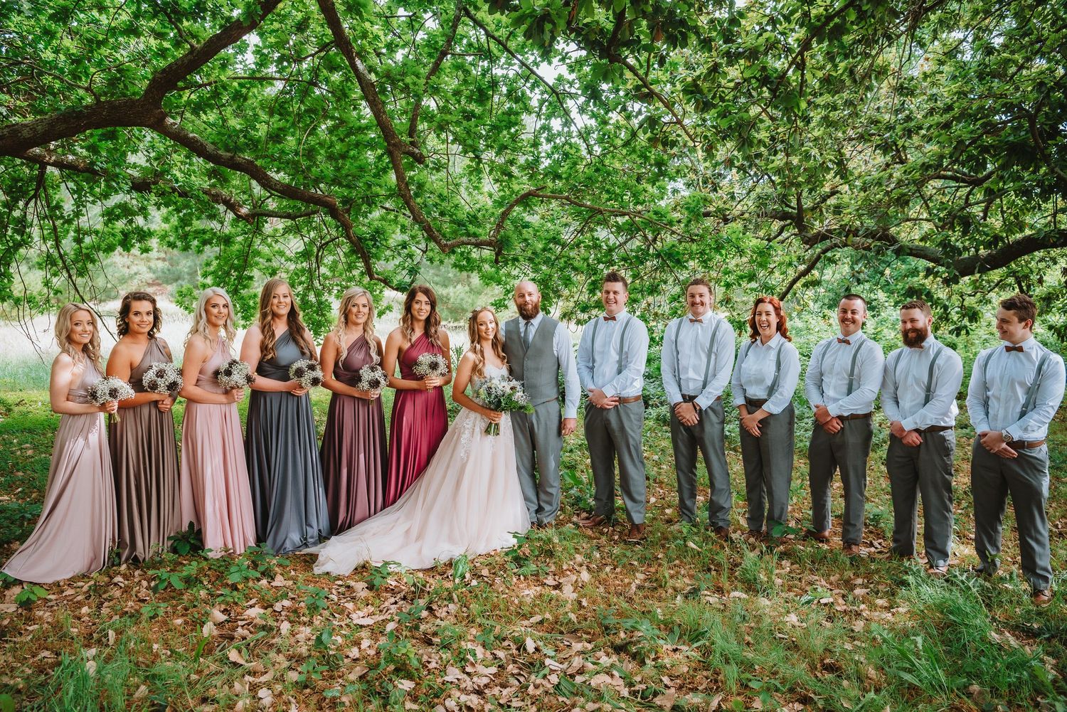 Wedding party poses outdoors in pink and gray attire beneath green trees during summer celebration.