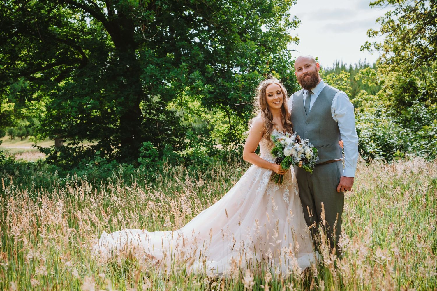 A couple stands together in a sunlit meadow with tall grass, the bride wearing a flowing lace dress and holding a bouquet.