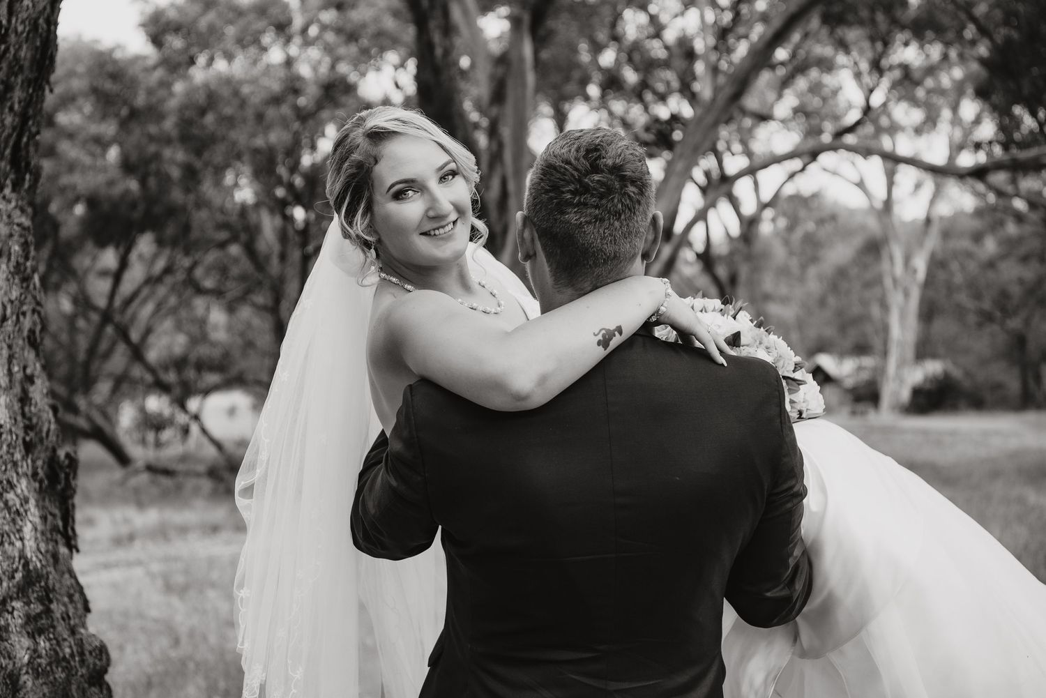 Black and white wedding photograph showing a tender moment between bride and groom embracing outdoors among trees.
