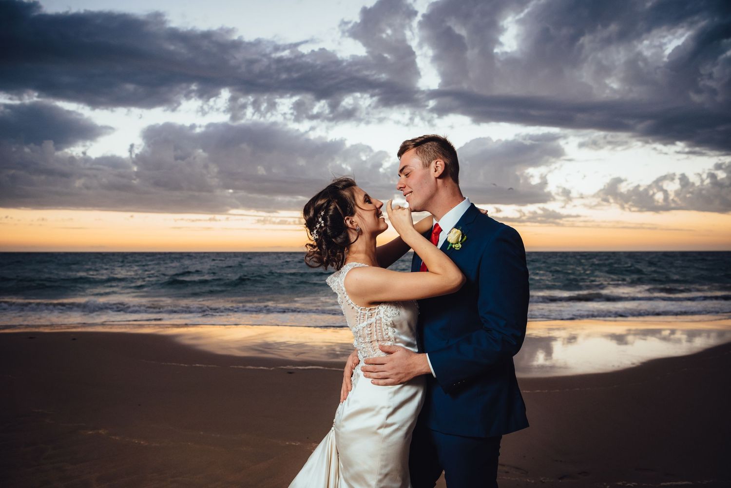 A couple embraces on a beach at sunset with dramatic storm clouds and ocean waves in the background.