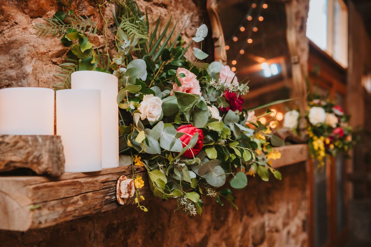 White candles and floral arrangement with pink roses and greenery on rustic wooden shelf with warm lighting in background.