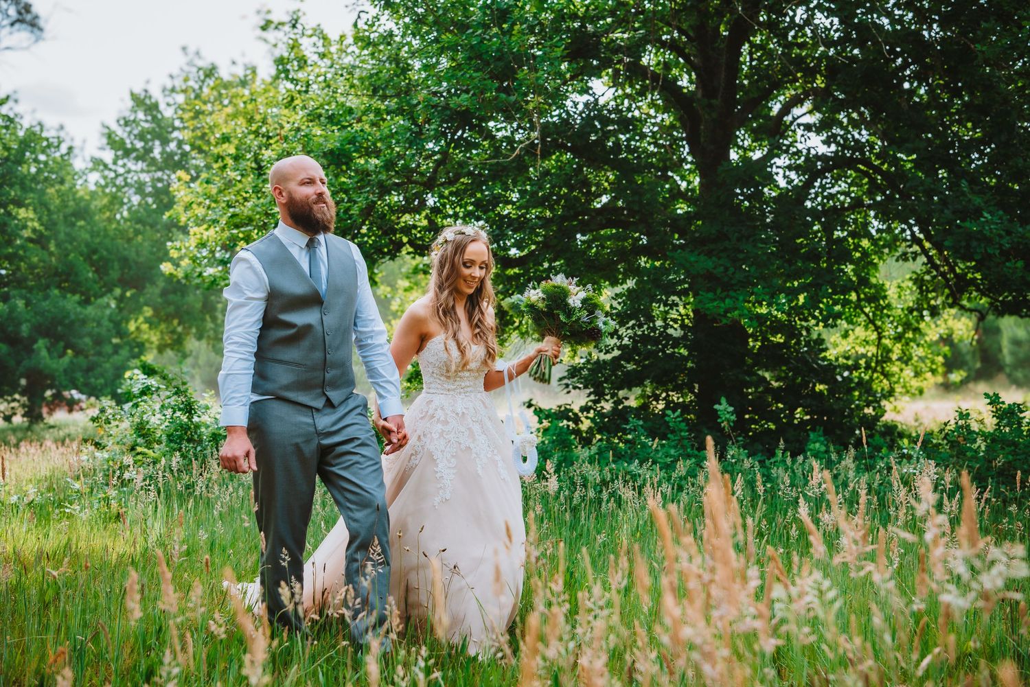A couple holds hands while walking through a lush summer meadow with tall grass and trees in the background.
