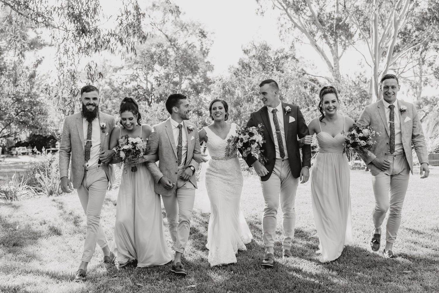 Elegant wedding party in light gray attire walking together outdoors with bouquets in hand during a joyful celebration.