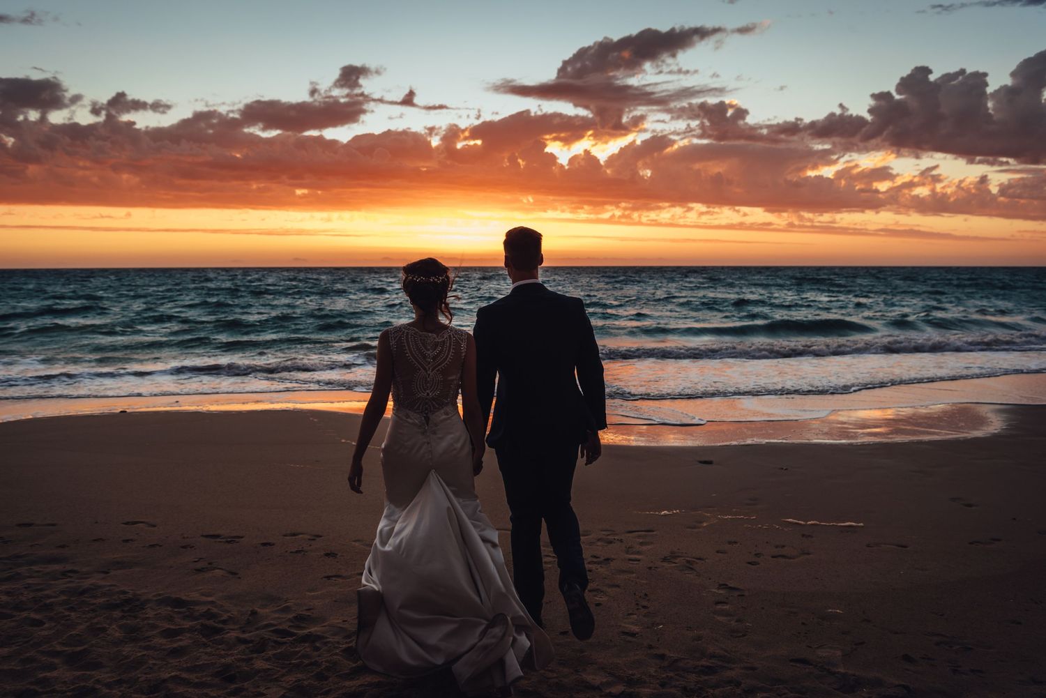 A couple in wedding attire walks along a beach at sunset with dramatic orange clouds over the ocean.