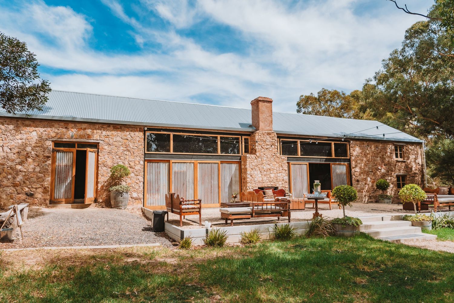 Modern stone farmhouse with large glass windows, timber deck and rustic chimney nestled among trees in rural setting.