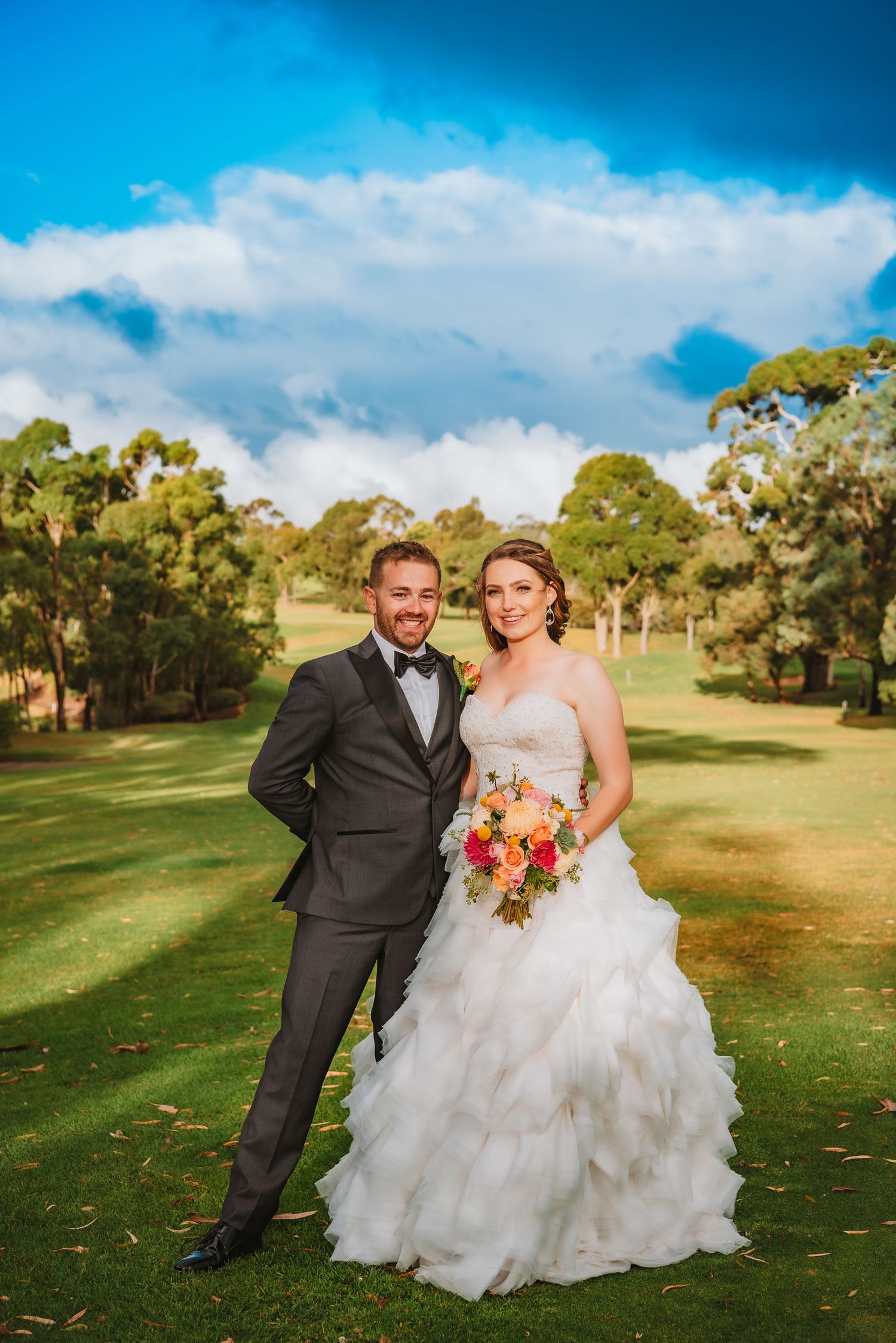 A couple stands together on a green golf course with white ruffled wedding dress and black tuxedo under blue sky.