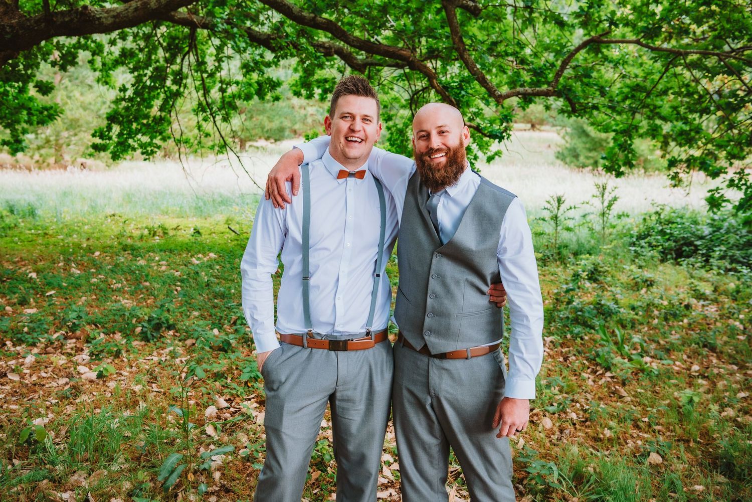 Two people wearing gray suits stand together smiling under tree branches in an outdoor garden setting.