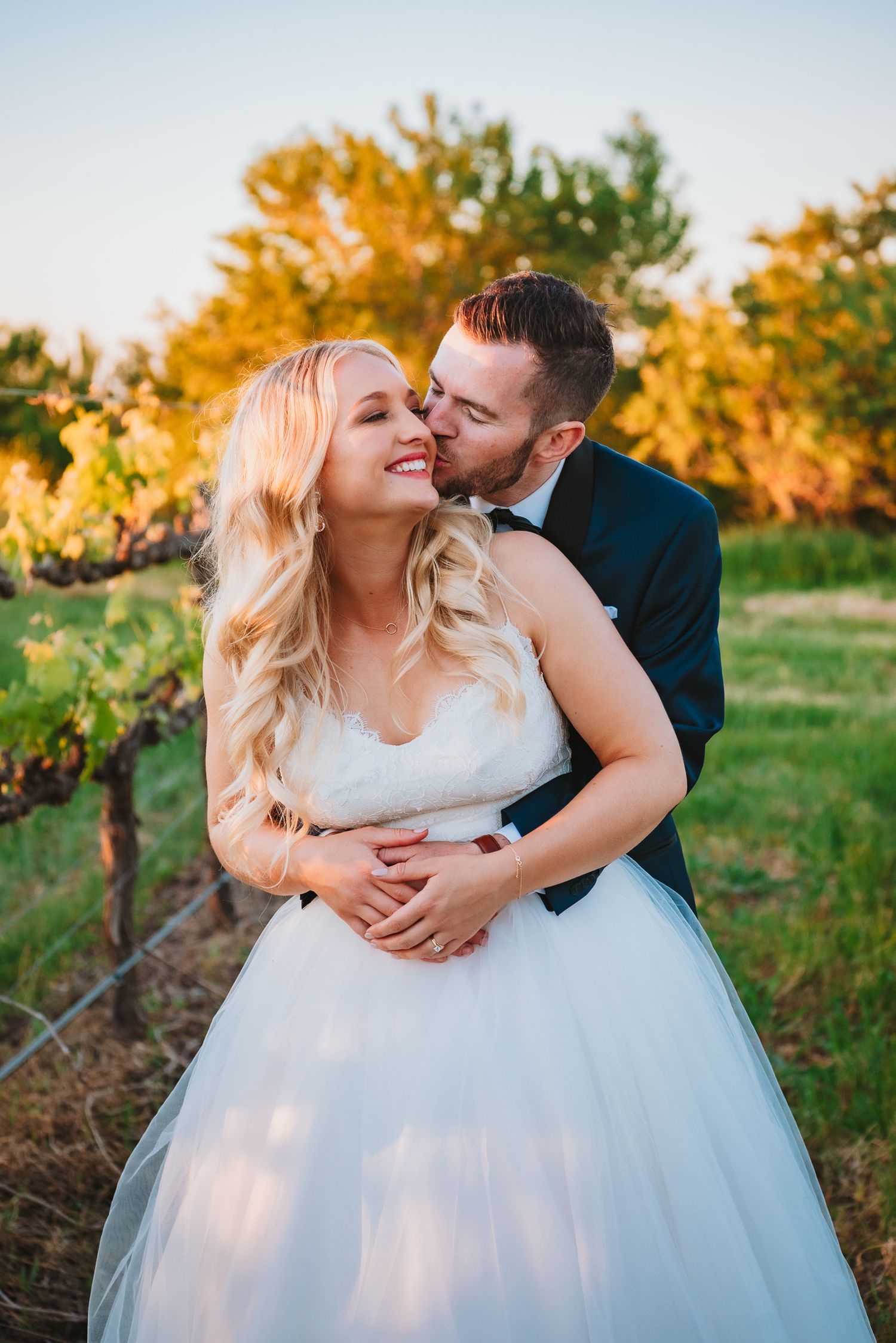 Romantic wedding couple embraces in vineyard during golden sunset light with elegant white gown and dark suit.