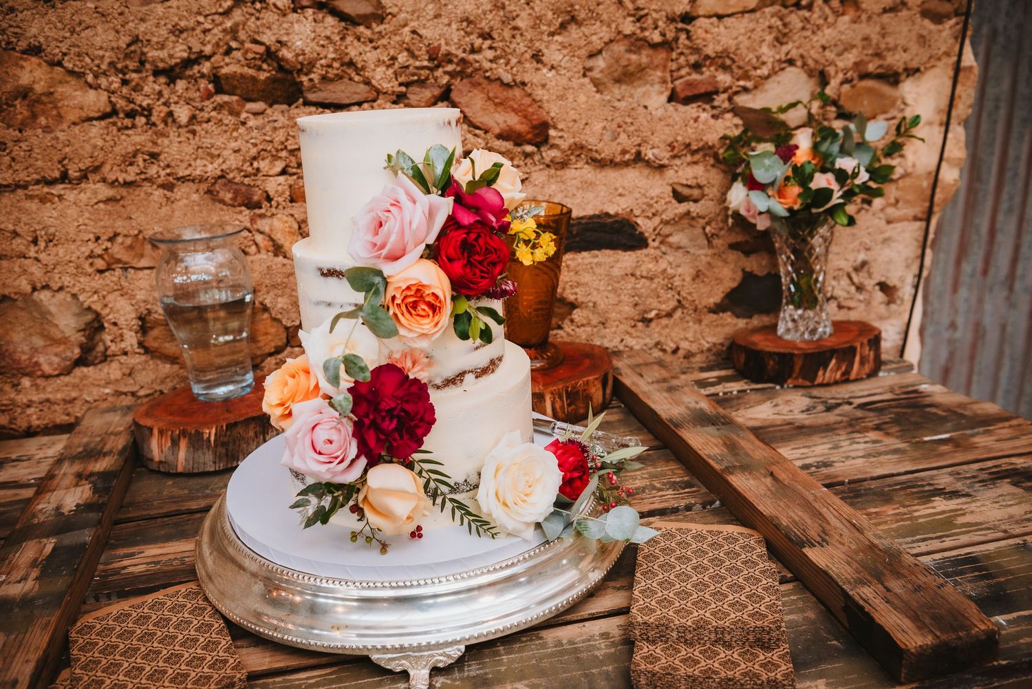 A rustic wedding cake decorated with pink, red and peach roses sits on a silver stand against a stone wall.