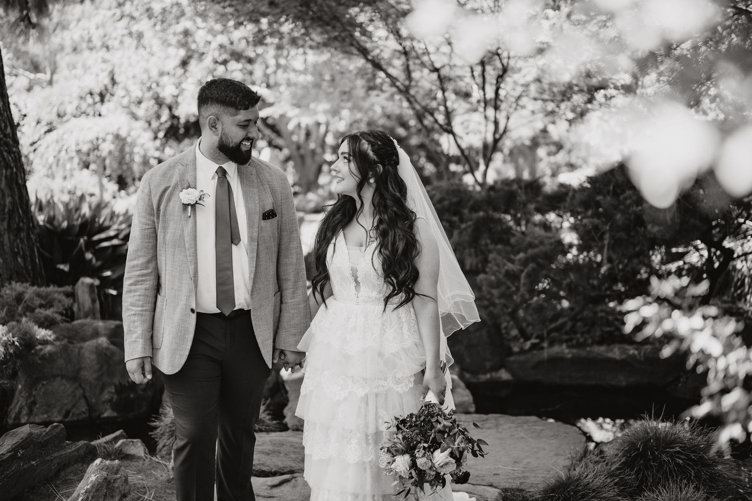 A romantic black and white wedding photo of a bride and groom smiling at each other outdoors amidst garden scenery.