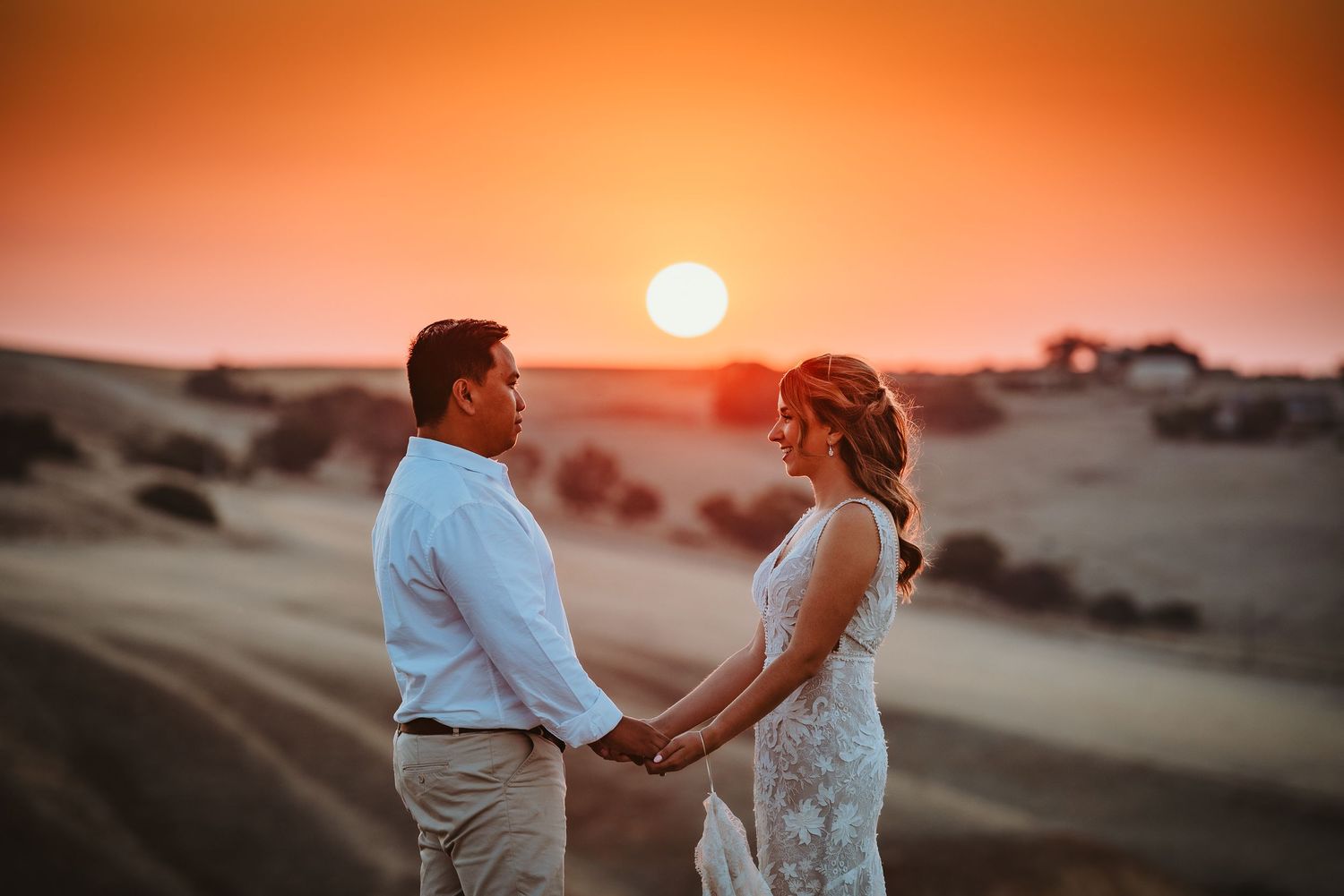 A couple holds hands in white attire against sand dunes at sunset with a glowing orange sky in the background.